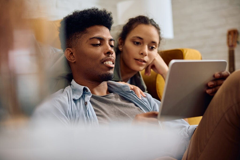 A couple sits close together on a couch, looking at a tablet screen and relaxing at home.