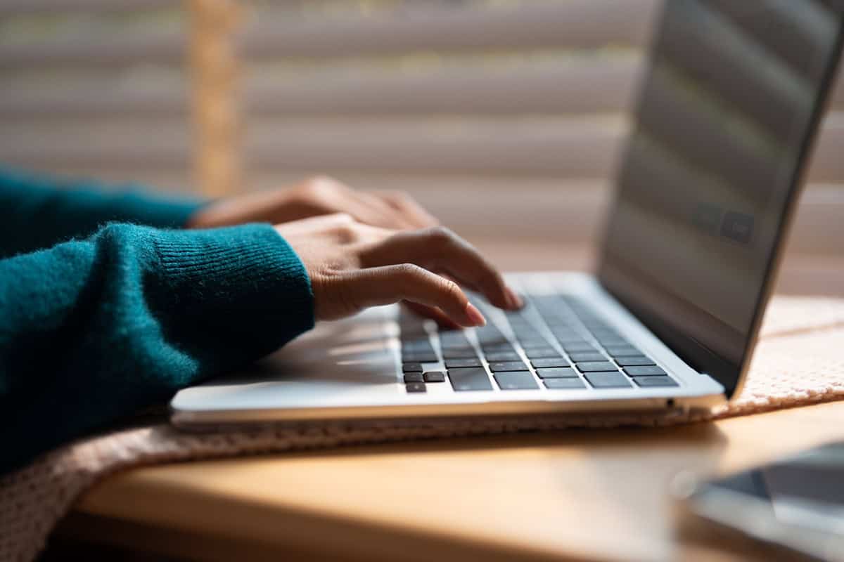 Close-up of hands typing on a laptop keyboard near a window with blinds.