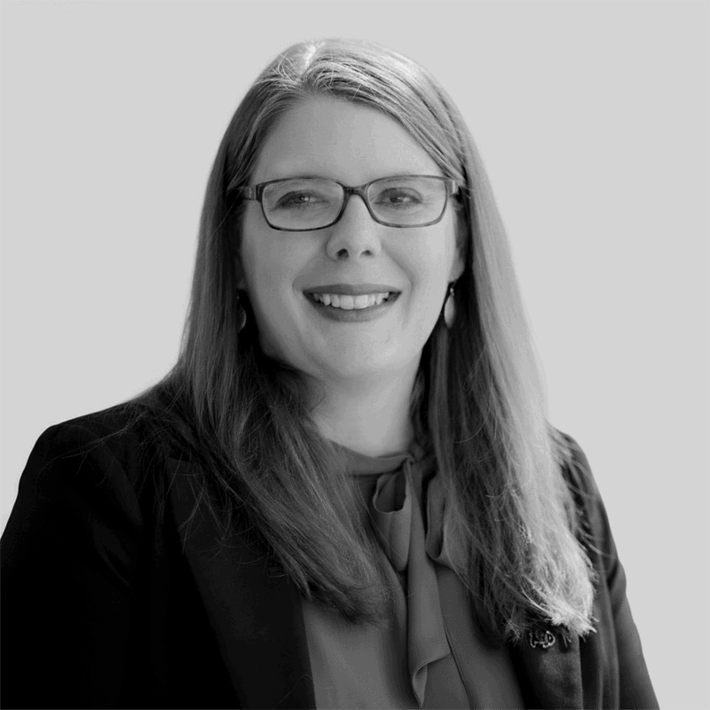 A woman with long hair and glasses, smiling, wearing a blazer and blouse, in a black and white photo.