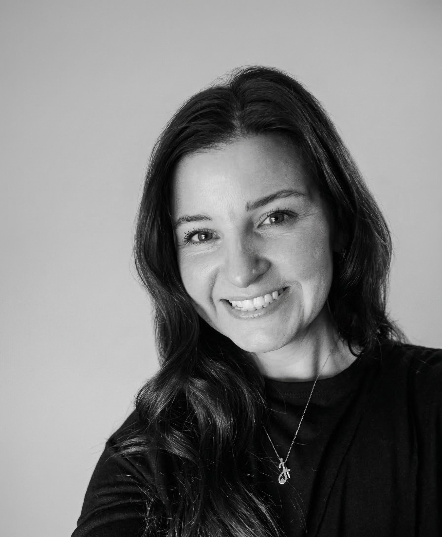 Smiling woman with long dark hair wearing a black top and necklace, against a plain background.