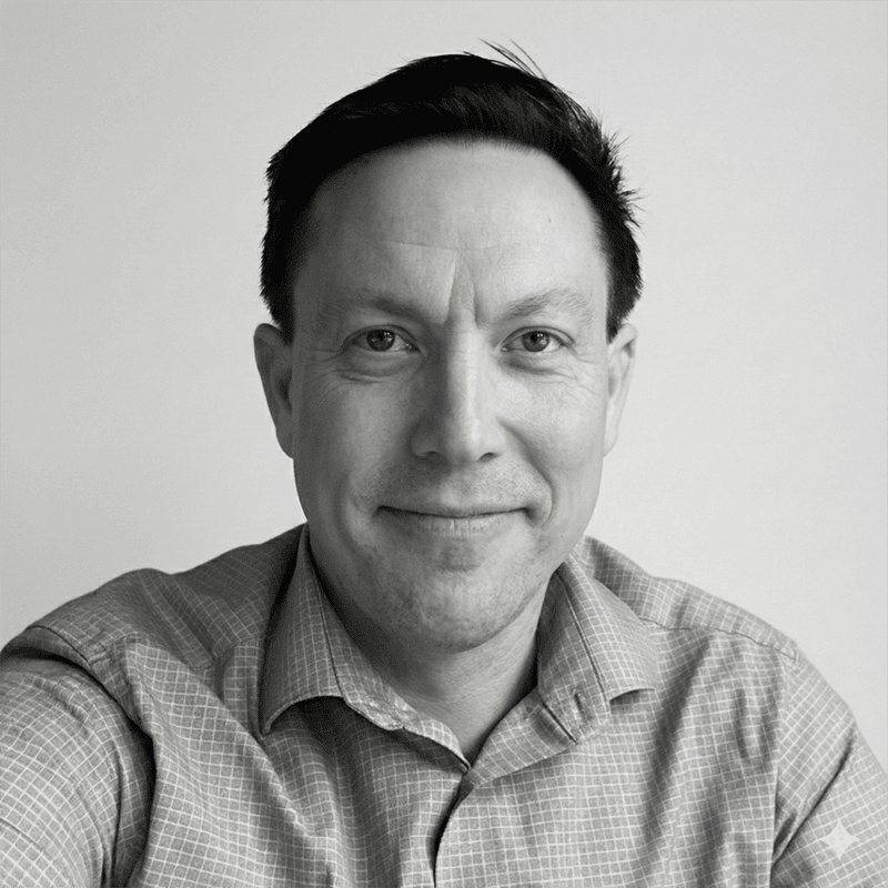 Man smiling at the camera, wearing a checked shirt, in a black-and-white portrait against a plain background.